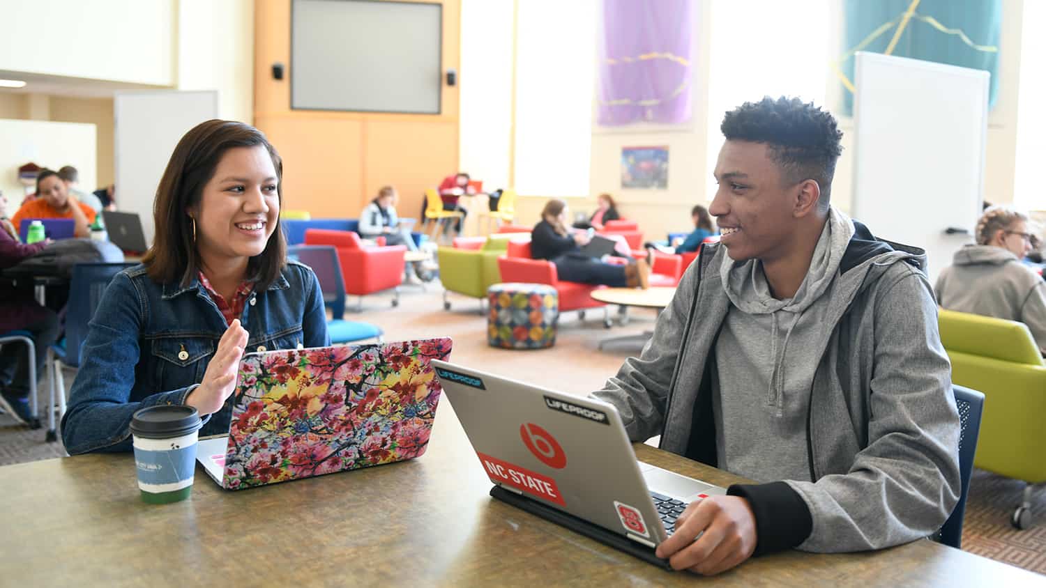 Two students talk while working on their laptops at a table in the Riddick Hall Hearth