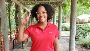 A female student makes the wolf ears sign while standing on a tree-lined sidewalk