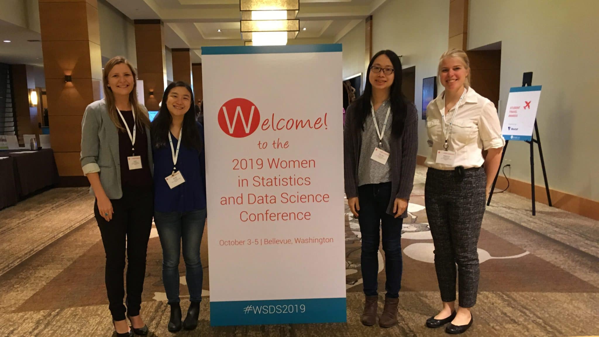 Four students stand on either side of a banner at the 2019 Women in Statistics and Data Science Conference