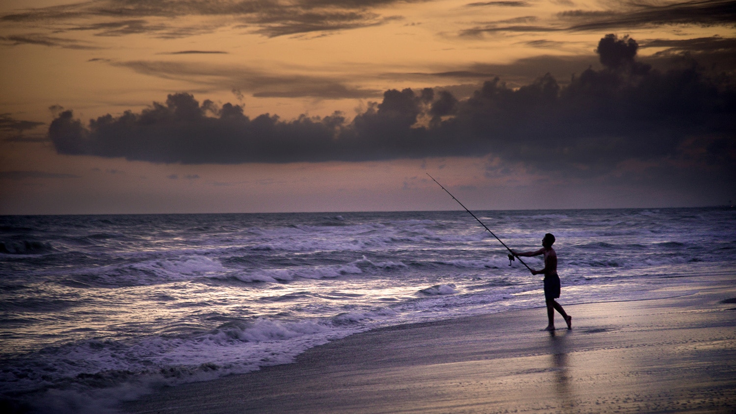 A fisherman on a beach casts his fishing rod into the ocean with the sun setting in the background