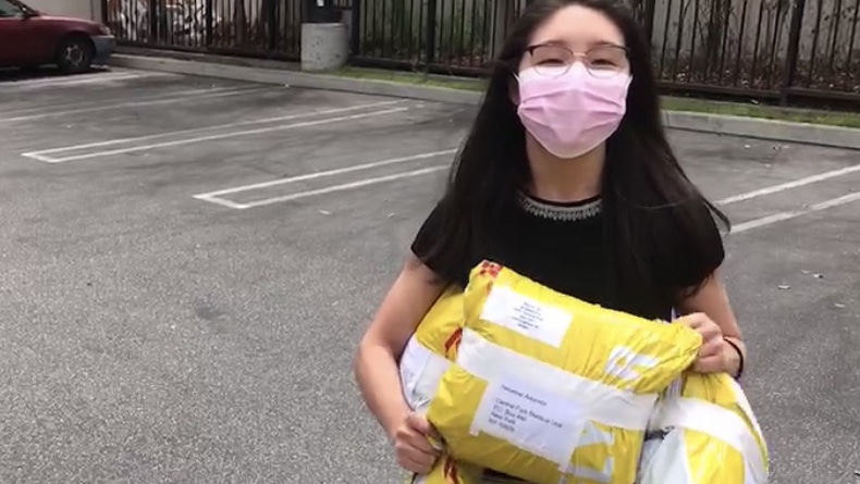 A woman stands in a parking lot wearing a face mask and carrying packages