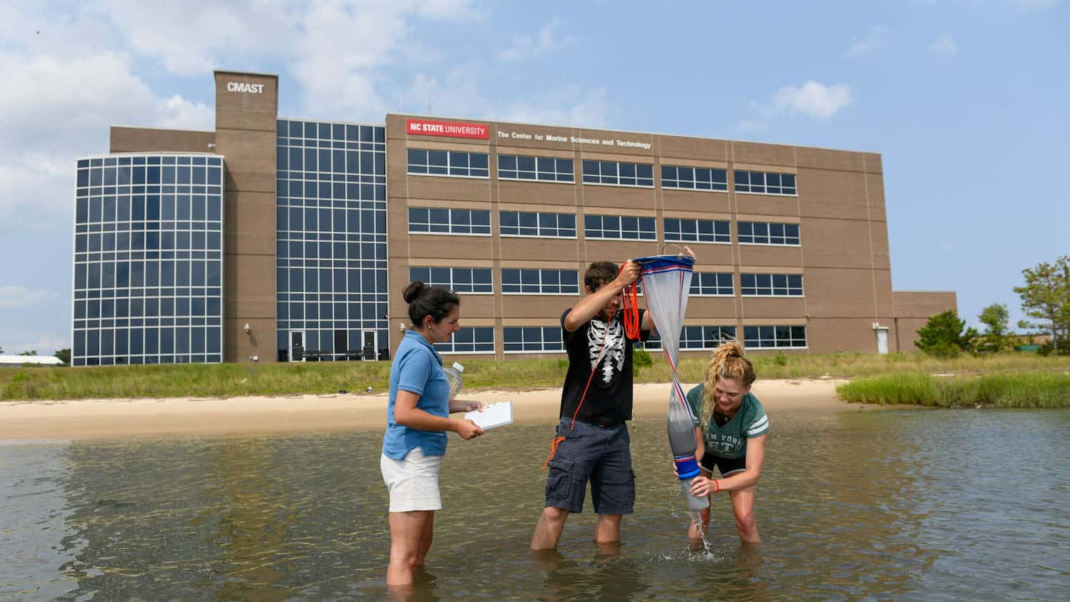 Researchers collect plankton in knee-deep water with the CMAST building in the background