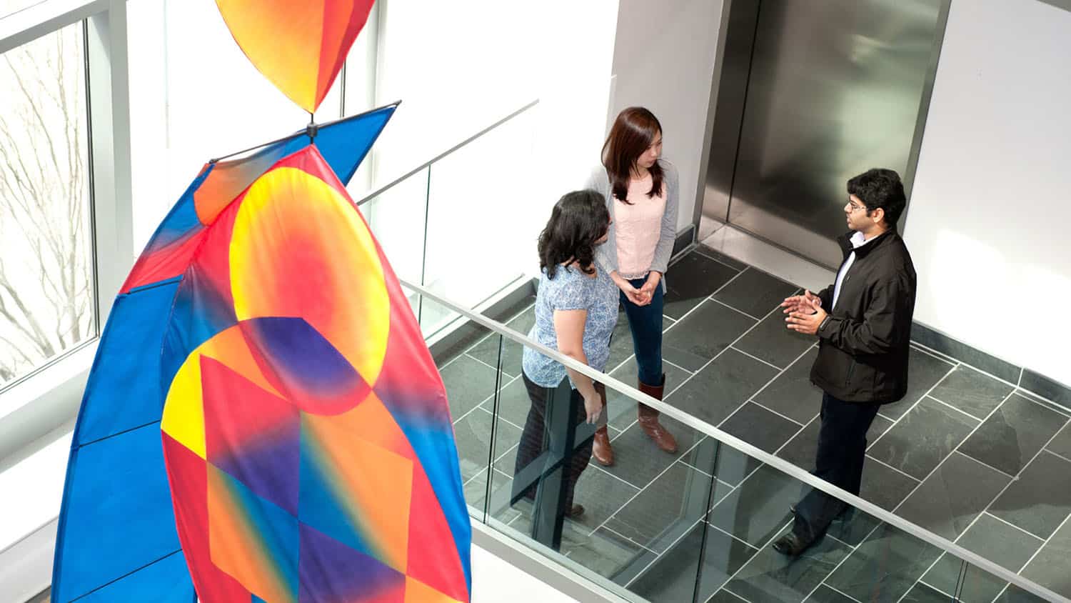 Three people talk in the Atrium of SAS Hall with a colorful art installation in the foreground