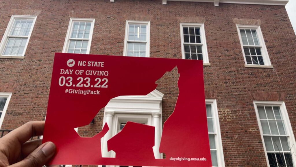 A Day of Giving postcard is held up on the NC State campus.