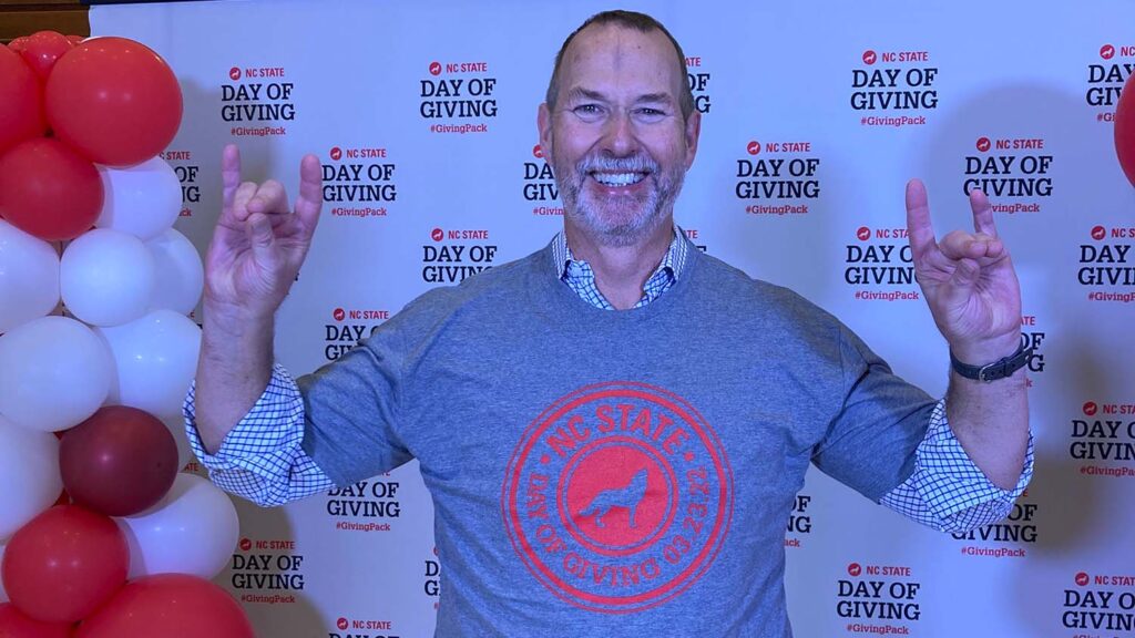 A man stands in front of an NC State Day of Giving background, making the Wolfpack sign with his hands. 