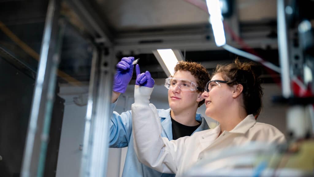 Adam Nahor holds up a microscope slide while Sierra Hunter points at it.