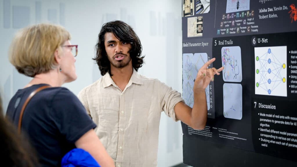 Aloha Das standing in front of his research poster, presenting his findings to someone.