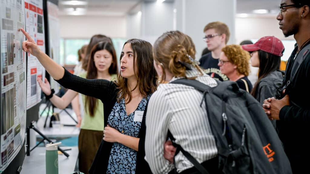 Katya Frazier, surrounded by people, points at her research poster.