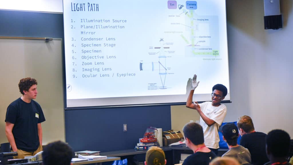 A student points to a slideshow as he presents in front of a class.