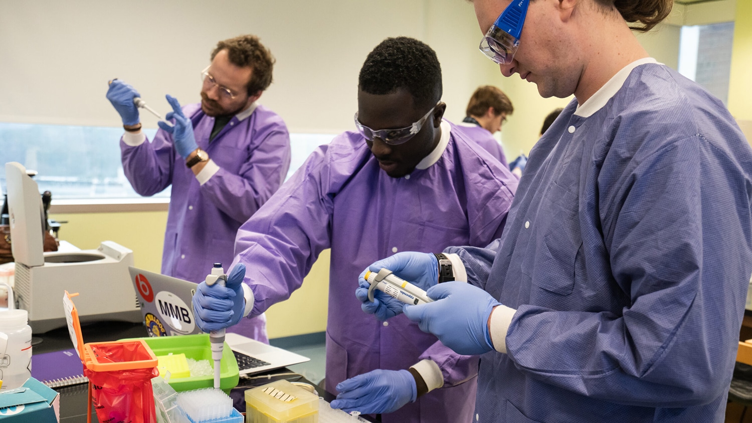 Researchers wearing lab coats and goggles work with pipettes